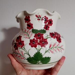 May include: A white ceramic vase with a ruffled rim, adorned with hand-painted red flowers, green leaves, and pink accents. The vase has a bulbous shape and a green base, perfect for floral arrangements.