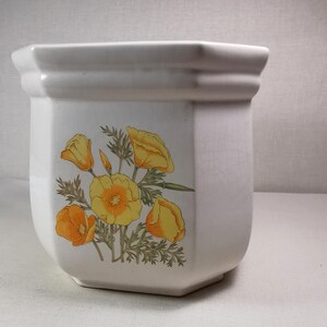 Pode incluir: Vaso de cerâmica branco com forma hexagonal e um design floral com papoulas da Califórnia amarelas e laranja.