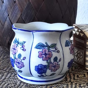 May include: A white ceramic planter with a scalloped rim and blue and pink floral design. The planter has a slightly rounded shape and a blue rim.
