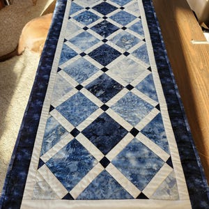 May include: A blue and white table runner with a diamond pattern. The runner has a dark blue border with a snowflake design.