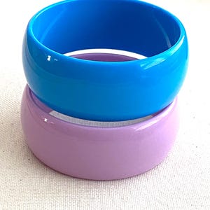 May include: Two smooth, wide bangle bracelets stacked vertically. The top bangle is a vibrant, solid bright blue, while the bottom bangle is a solid, soft pastel lavender. Both bracelets feature a glossy, reflective finish and a rounded, thick profile.