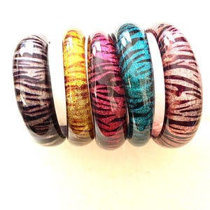 May include: Five colorful bangle bracelets with a zebra print design. The bracelets are made of glass and have a shiny finish. The colors of the bracelets are purple, gold, pink, teal, and maroon.