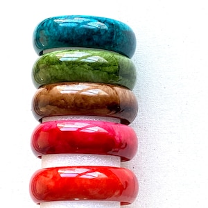 May include: A stack of five colorful bangles in shades of teal, green, brown, pink, and red. The glossy, rounded bracelets are stacked vertically, showcasing their vibrant hues and smooth surfaces. The bangles are approximately 2.5 inches in diameter.