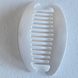 May include: White plastic hair comb with a curved shape and a row of teeth. The comb is designed to hold hair in place.