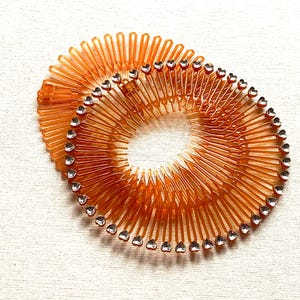 May include: Two orange hair combs with a circular design. Each comb features a series of thin, curved teeth and is adorned with small, clear heart-shaped embellishments around the outer edge. The combs are set against a white background.
