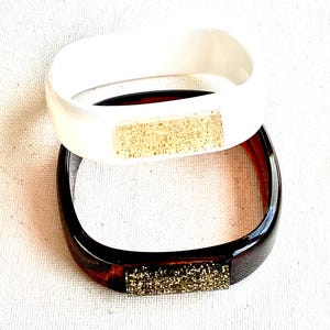 May include: Two plastic bangles with a square shape. The top bangle is white with a gold glitter accent. The bottom bangle is brown with a gold glitter accent.