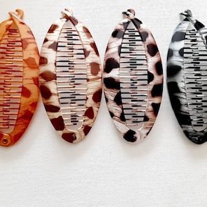 May include: Four brown, black, and white patterned hair clips with a comb design. The clips are shaped like a long oval.