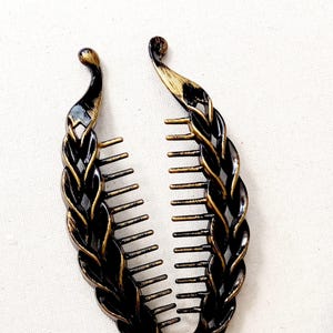 May include: A banana hair clip featuring a curved, interlocking comb design. The accessory has a dark black base with metallic gold-toned highlights brushed along the raised, braided-style texture of the outer edges and the individual teeth.