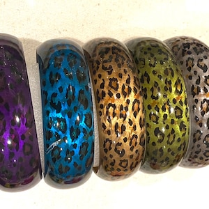 May include: Five colorful bangles with a leopard print design. The bangles are in shades of purple, blue, gold, green, and silver. The bracelets are made of a glossy material and are displayed on a white surface.