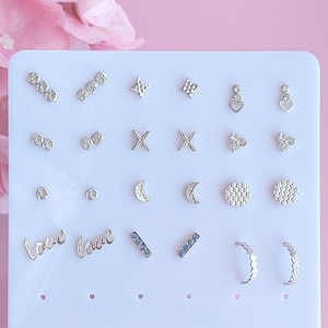 Può includere: Una scheda di esposizione bianca con 18 paia di orecchini d'argento. Gli orecchini sono disposti in file e includono vari design, come stelle, cuori, lune e la parola "love". La scheda ha la parola "JEMCOAST" stampata in basso.