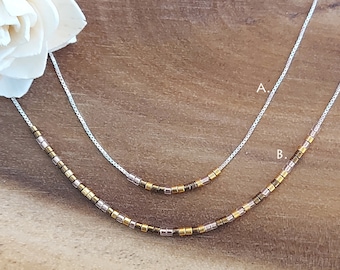 May include: Two delicate necklaces with silver chains and small gold, bronze, and pink beads. The necklaces are layered on a wooden surface. The top necklace is labeled "A" and the bottom necklace is labeled "B".