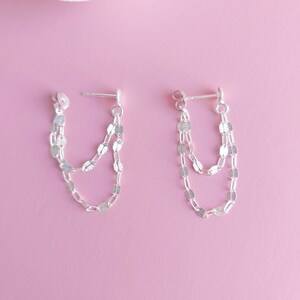 May include: A pair of silver earrings with a delicate chain design. The earrings feature two layers of chains with small, oval-shaped links. The chains dangle from a small, silver post.
