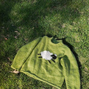 May include: A lime green long-sleeve knit sweater featuring a crew neckline and a ribbed collar. The center of the sweater displays a knitted graphic of a white sheep with a black head and black legs, crafted with a textured yarn finish.