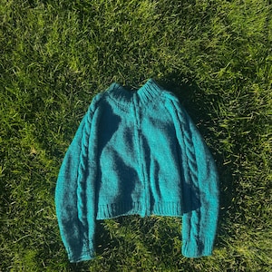 Könnte beinhalten: Ein langärmeliger Strickpullover in Petrol mit Stehkragen, gerippten Bündchen und einem gerippten Saum. Der Pullover ist mit einem vertikalen Zopfstrickmuster entlang der Ärmel und einer mittigen Naht auf der Vorderseite gearbeitet.