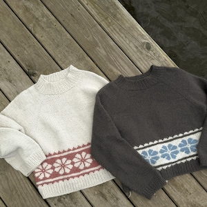 Könnte beinhalten: Zwei langärmelige Strickpullover mit gerippten Ausschnitten und Bündchen. Der linke Pullover ist cremefarben mit einem horizontalen Band aus rosa Blumenmustern. Der rechte Pullover ist dunkelgrau mit einem Band aus hellblauen Blumenmustern.