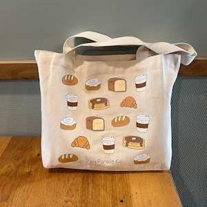 Puede incluir: Una bolsa de lona blanca con un asa marrón. La bolsa tiene una variedad de ilustraciones de alimentos para el desayuno, que incluyen pan, pasteles, café y panqueques. El texto "Yay Parade Co." está impreso en la parte inferior de la bolsa.