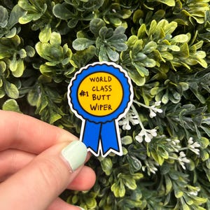 May include: A sticker featuring a blue ribbon design with the text "WORLD CLASS BUTT WIPER" in black on a yellow circle. The sticker also has the number "#1".