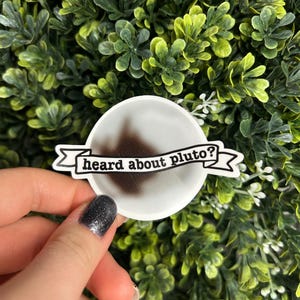 May include: A white round sticker with a black banner that says "heard about pluto?" The sticker is being held by a hand with black nail polish.