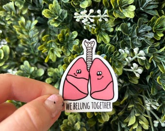 Könnte beinhalten: Ein rosa-weißer Aufkleber mit einer Cartoon-Abbildung von zwei Lungen mit lächelnden Gesichtern. Der Text "We Belong Together" ist unter den Lungen gedruckt.