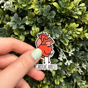 Puede incluir: Un corazón rojo de dibujos animados con una expresión preocupada sostiene un desfibrilador. El texto "Cardiac Arrest" está debajo del corazón.