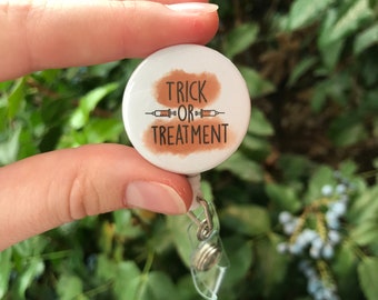 May include: White button with a brown watercolour background and the text "Trick or Treatment" with two syringes on either side of the word "or". The button is attached to a clear plastic retractable badge holder.