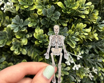 May include: A white skeleton sticker with brown lines outlining the bones. The sticker is held in a hand with light green nail polish.
