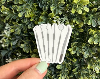 Puede incluir: Una pegatina blanca con un conjunto de seis herramientas dentales, que incluyen un espejo, una sonda y un raspador. Las herramientas están dispuestas verticalmente y están delineadas en gris.