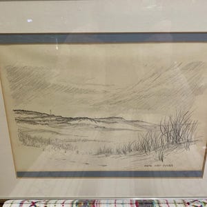 Puede incluir: Un boceto a lápiz enmarcado de un paisaje costero. La obra representa dunas, hierbas altas y un cielo nublado. El boceto está firmado "CAPE COD DUNES" en la esquina inferior derecha. El marco es plateado con un paspartú azul.