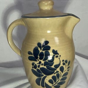 Puede incluir: Una jarra de cerámica de color beige con un diseño floral azul y una tapa. La jarra tiene un asa y un pico.