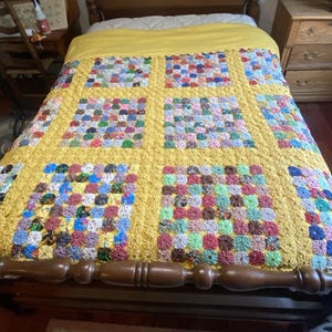 Może przedstawiać: Ręcznie robiona kołdra z żółtym tłem i kolorowymi kwiatowymi łatkami. Kołdra znajduje się na drewnianej ramie łóżka. Wzór kołdry zawiera kwadraty w różnych kolorach, w tym czerwonym, niebieskim, zielonym i różowym.