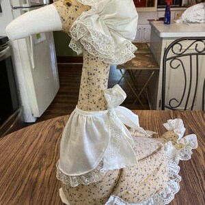 May include: A stuffed fabric goose figurine featuring a beige body with a small blue and brown floral pattern. The goose wears a white bonnet with lace trim, a white fabric collar with a bow, and white lace trim along its tail feathers.