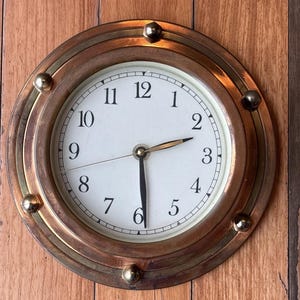 May include: Round clock with a white face and black numerals, set in a bronze and brass frame. The clock has a classic design with a second hand and is mounted on a wooden surface.