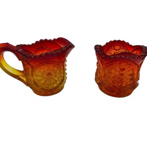 Könnte beinhalten: Zweiteiliges Set aus antikem Karnevalsglas. Das Set beinhaltet einen kleinen Krug mit Henkel und einen passenden Becher. Das Glas hat eine lebendige orange-gelbe Irisierung mit einem detaillierten Muster.