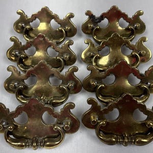 以下が含まれることがあります： 装飾的なデザインの、真鍮製のアンティーク引き出しプルが8つ。各プルは、湾曲したハンドルと、2つの円形の開口部を備えた詳細なバックプレートを備えています。真鍮は暖かく、古びた風合いで、ヴィンテージまたは再生されたハードウェアを示唆しています。