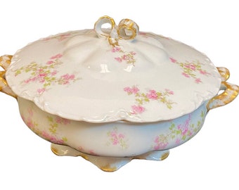 May include: A white porcelain tureen with a scalloped edge, featuring a delicate pink floral pattern and green foliage. The lid has a decorative gold-trimmed handle, and the base includes gold-painted accents on the handles and feet.
