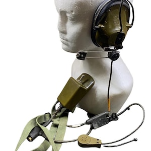 May include: An olive-green military-style headset with a microphone, ear cups, and a headband. The headset is attached to a strap and a rectangular control box. The headset is displayed on a white mannequin head.