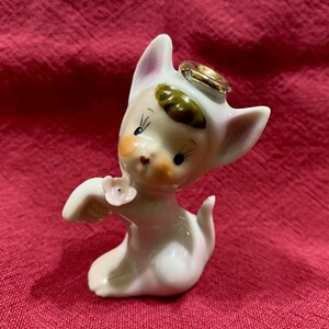 May include: A vintage ceramic figurine of a cat-like creature with a white body, pink-lined ears, and a gold halo. The figurine has a painted face with rosy cheeks and holds a small pink flower. The figure is kneeling on a red surface.