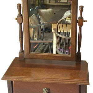 Può includere: Uno specchio da toeletta in legno vintage con base rettangolare e cassetto. Lo specchio è incorniciato in una forma arcuata curva. Il cassetto presenta una maniglia a testa di leone in ottone. Lo specchio è sostenuto da due montanti in legno regolabili.