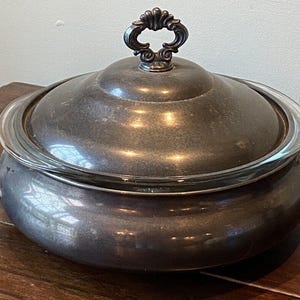 Könnte beinhalten: Silberne Servierplatte mit Glasdeckel und verziertem Griff. Die Platte hat eine runde Form und eine glatte, reflektierende Oberfläche.