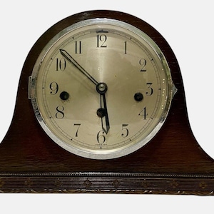 May include: A vintage wooden mantel clock with a silver-colored face and black hands. The clock has a dark brown, curved wooden frame with decorative carvings along the base. The clock face displays the numbers 1 through 12.