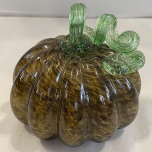 Puede incluir: Una calabaza decorativa de vidrio con cuerpo marrón y tallo verde. La calabaza tiene una textura acanalada y un patrón arremolinado. El tallo es de un verde más claro y tiene un diseño rizado. Este artículo con temática otoñal es adecuado para la decoración del hogar.