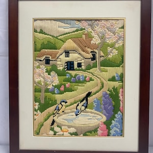 May include: A framed needlepoint embroidery depicting a cottage in a green landscape with a winding path, a bird bath, and two birds. The cottage has a brown roof and white walls with a blue door. The landscape is filled with flowers in shades of pink, purple, and yellow.