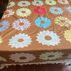 May include: A brown bedspread featuring large, colourful flower appliqués. The flowers are in shades of pink, blue, red, and yellow, with white centres. The top of the bed has a blue and yellow pineapple pattern.