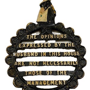 Puede incluir: Salvamanteles de hierro fundido negro y dorado con una figura de mujer en la parte superior. El salvamanteles tiene un diseño circular con el texto "THE OPINIONS EXPRESSED BY THE HUSBAND IN THIS HOUSE ARE NOT NECESSARILY THOSE OF THE MANAGEMENT."
