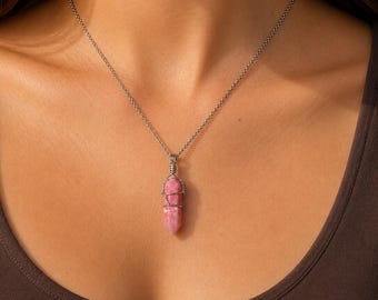 May include: A necklace featuring a polished, cylindrical pink crystal pendant wrapped in silver-toned wire. The pendant hangs from a delicate, thin silver-toned metal chain that rests against the collarbone, showcasing a smooth, stone texture.