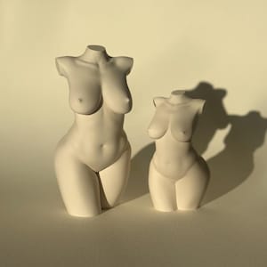 May include: Two white ceramic sculptures of the female torso. The sculptures are in different sizes, with the larger one on the left and the smaller one on the right.
