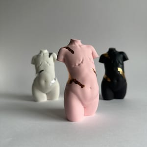 Könnte beinhalten: Drei Keramik-Torso-Skulpturen, jede in einer anderen Farbe: weiß, rosa und schwarz. Jede Skulptur hat goldene Akzente auf dem Torso.