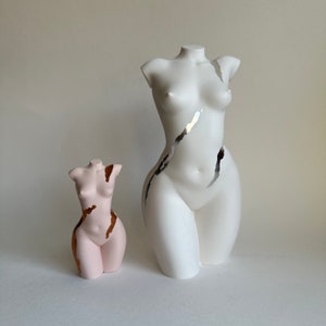 May include: Two decorative torso sculptures. One is white with silver and black accents, the other is pink with gold accents. Both sculptures are headless and depict the female form.
