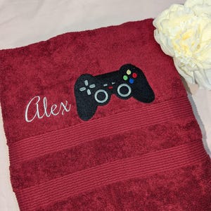 May include: A red bath towel with a black embroidered video game controller and the name "Alex" in white embroidery.