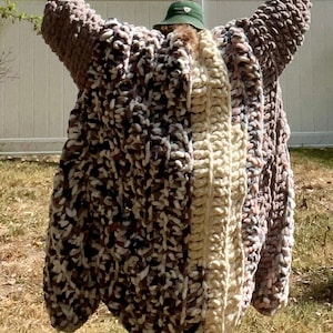 May include: A person wearing a long, oversized, crocheted cardigan with a white stripe down the center. The cardigan is made of a chunky yarn in shades of brown, pink, and white. The person is wearing a green hat and is standing in a grassy area.
